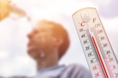 Adam aşırı sıcakta kafasına su döktü. Yaz günündeki termometre arka planda güneş olan yüksek sıcaklık derecesini gösterir veya gösterir.
