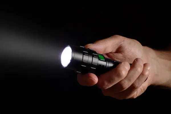Male hand holding a led flashlight with a wide white beam on a black background, leaving the right side of the frame.