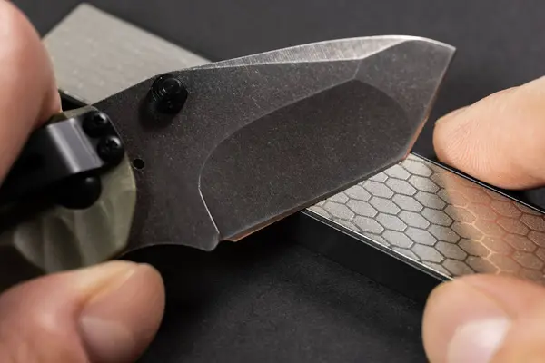 stock image Man sharpens a knife using a whetstone on a dark table.