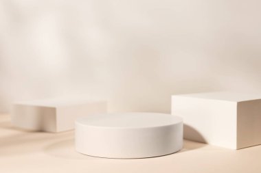 Bej arka planda üç beyaz podyum ve ağaç yapraklarının gölgeleriyle boş bir sahne. Kozmetik ürün ve ambalaj modelleme sunumu için kaldırım taşı