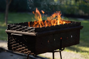 Yanan odundan çıkan portakal ateşi dilleri. Közlerin, yeşil çimlerin bulanık arka planında ızgarada barbekü hazırlığı için hazırlanması..