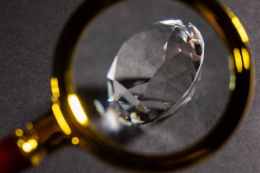 Karanlık bir arkaplan önünde elmaslar, yerde yansımalar. Cımbızla tutulan parlak kesilmiş elmas..