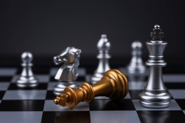 Gümüş kral satranç taşı, satranç tahtasındaki düşen altın kral taşının üzerinde muzaffer duruyor. Strateji, zafer ve liderlik kavramı.