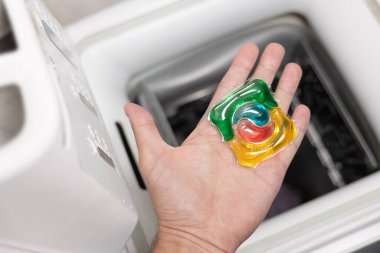 Çamaşır deterjanı kapsülünü çamaşır makinesine koyan adam, yakın plan. Kapsülde renkli çamaşır ekosu jeli. Çamaşır yıkıyorum. Yıkama ve temizleme kavramı