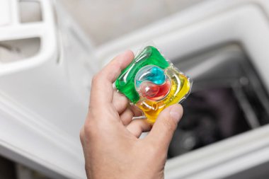 Erkek eli, çamaşır makinesinin arka planında beyaz çamaşırlı bir çamaşır kapsülü tutuyor..