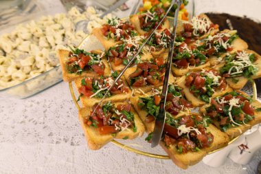 Taze doğranmış domates, şifalı otlar ve kıtır ekmeğin üzerinde doğranmış peynirle kaplı güzel bir ev yapımı bruschetta tabağı. Tabak, dantel bir masa örtüsünün üzerinde zarif bir altın sehpada sunuluyor. Sıcak ve davetkar bir ortam yaratıyor.