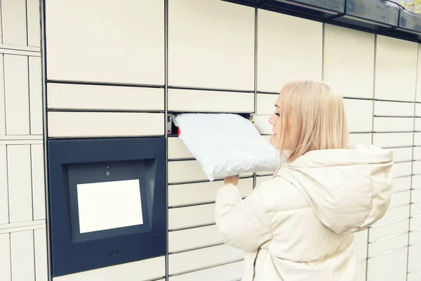 Blonde woman put the parcel in the self-service mail terminal. Parcel delivery machine.