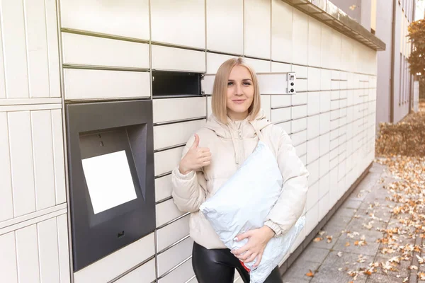 A blonde woman holds a parcel and a finger of approval near the self-service mail terminal. Parcel delivery machine.