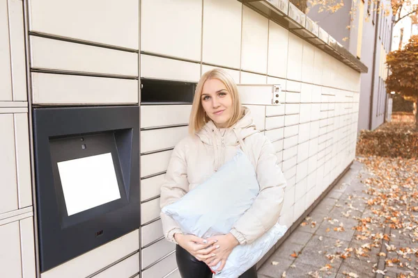 Blonde woman hold the parcel near self-service mail terminal. Parcel delivery machine.
