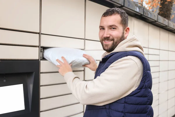 Happy bearded man put the parcel in the self-service mail terminal. Parcel delivery machine.