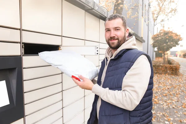 Happy bearded man put the parcel in the self-service mail terminal. Parcel delivery machine.