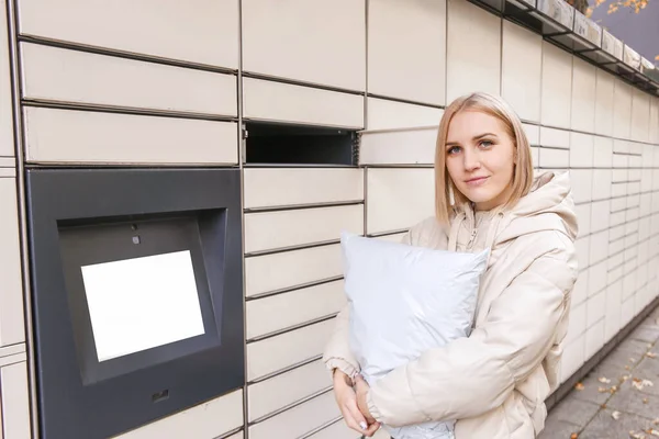 Blonde woman hold the parcel near self-service mail terminal. Parcel delivery machine.
