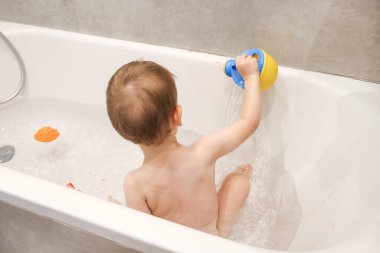 Çocuk banyoda bir su bidonuyla, kalemlerle, banyoda oyunlar, banyo yapan çocuklarla oynuyor.