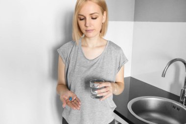 Sarışın kız elinde haplarla mutfakta su içiyor. Vitaminler. Hastalık. Doğum kontrol hapı. Sağlıksız. Tedavi