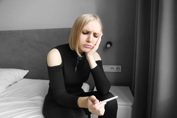 sad blonde girl sitting in the bedroom and looking at the pregnancy test