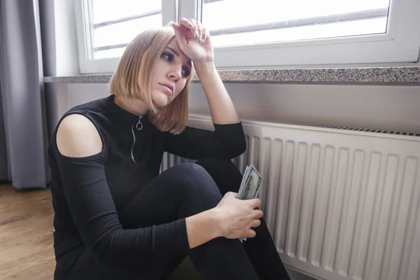 Blonde woman sits near the heating radiator and hold money. Rising utility bills. A crisis. Expensive heating bill