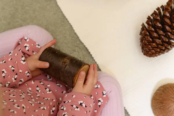 Yukarıdan bakıldığında, pembe desenli uzun kollu bir gömlek ve açık mor taytlar giyen küçük, silindirik tahta bir kütük tutan bir çocuğun elleri görülüyor. Çocuk beyaz desenli bir yüzeye yakın açık gri bir halıda oturuyor, muhtemelen bir hayvan.
