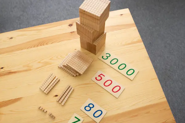 Montessori matematik materyallerinin yakın plan bir fotoğrafı, açık tahta bir masa üzerine dizilmiş. Malzemeler arasında binlerce küplük bir kule, bir araya toplanmış yüzlerce kare, birden fazla onluk çubuk ve birkaç birim boncuk yer alıyor.