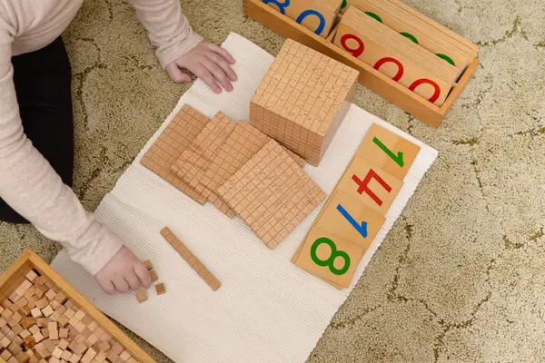 Yüksek açılı bir çekim, bej bir halıya yerleştirilmiş beyaz bir paspasın üzerinde Montessori matematik materyalleriyle etkileşen bir çocuğun ellerini gösteriyor. Uzun kollu açık bej gömlek ve siyah pantolon giyen çocuk küçük tahta küpleri düzenliyor. Dağılın.