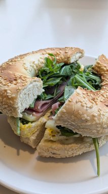 Taze roka, jambon, yumurta ve peynirle dolu susamlı bagel sandviçi resmi, rahat bir kafede yeşil bir tabakta servis edilir. Simit, brunch, dışarıda yemek, yemek, sandviç kavramı.