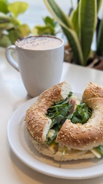 Taze roka, jambon, yumurta ve peynirle dolu susamlı bagel sandviçi resmi, rahat bir kafede yeşil bir tabakta servis edilir. Simit, brunch, dışarıda yemek, yemek, sandviç kavramı.