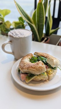 Taze roka, jambon, yumurta ve peynirle dolu susamlı bagel sandviçi resmi, rahat bir kafede yeşil bir tabakta servis edilir. Simit, brunch, dışarıda yemek, yemek, sandviç kavramı.