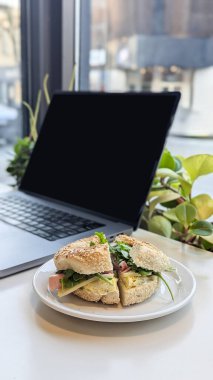 Kafe ortamında roka, jambon, yumurta, peynir ve susamlı simit sandviçi. Eş çalışma, brunch, simit, sandviç, üretkenlik, gevşek yemek, modern çalışma alanı kültürü kavramı.