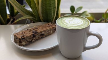 Kupada kalp şeklinde köpük sanatı olan bir kibrit latte, tabağında protein çubuğu olan yeşil bir masaya konur. Kibrit, kalp, latte, fincan, köpük, içecek, yeşil, bitki, latte sanat deseni kavramı.