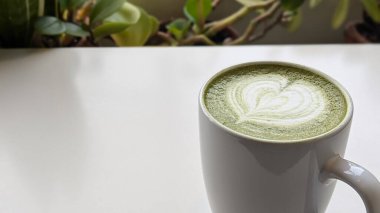 Beyaz seramik bardakta, kalp şeklinde köpük sanatı olan bir kibrit latte, yeşil bir masanın üzerine yerleştirilmiş. Kibrit, kalp, latte, fincan, köpük, içecek, masa, yeşil, beyaz, bitki ve desen kavramı.