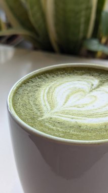 Beyaz seramik bardakta, kalp şeklinde köpük sanatı olan bir kibrit latte, yeşil bir masanın üzerine yerleştirilmiş. Kibrit, kalp, latte, fincan, köpük, içecek, masa, yeşil, beyaz, bitki ve desen kavramı.