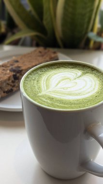 Kupada kalp şeklinde köpük sanatı olan bir kibrit latte, tabağında protein çubuğu olan yeşil bir masaya konur. Kibrit, kalp, latte, fincan, köpük, içecek, yeşil, bitki, latte sanat deseni kavramı.