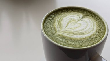 Beyaz seramik bardakta, kalp şeklinde köpük sanatı olan bir kibrit latte, yeşil bir masanın üzerine yerleştirilmiş. Kibrit, kalp, latte, fincan, köpük, içecek, masa, yeşil, beyaz, desen kavramı.