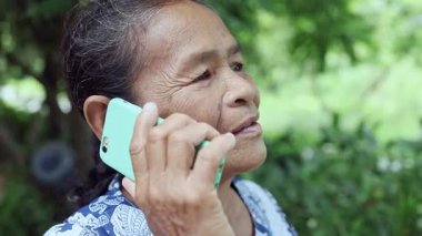 Mutlu Asyalı olgun kadın bahçede mutlu bir şekilde gülümsüyor, ailesiyle cep telefonunda konuşuyor, Tayland 'lı yaşlı büyükanne kablosuz teknoloji ve dijital cihazla dışarıda akıllı bir yaşam tarzının tadını çıkarıyor.
