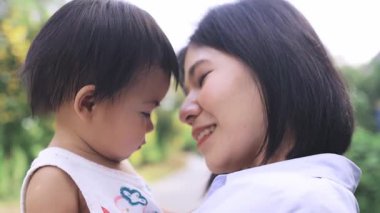 Asyalı bir anne ve bebeğinin açık havada gülümseyip burunlarına dokunduğu mutlu bir an. Saf mutluluk, sevgi ve aile bağının sıcaklığını yakalıyorlar. Günlük aile hayatında neşe.