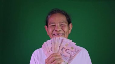 Mutlu Asyalı kadın elinde büyük bir gülümsemeyle Tayland baht banknotları tutuyor, finansal başarı kavramı, şans ve emeklilik sevinci krom anahtar yeşil ekran arka planında, büyükanne mutlu bir şekilde parasıyla