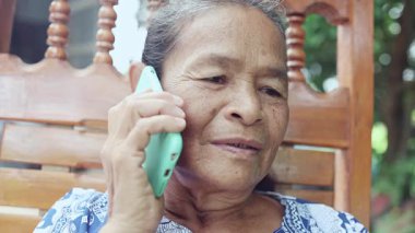 Mutlu Asyalı olgun kadın bahçede mutlu bir şekilde gülümsüyor, ailesiyle cep telefonunda konuşuyor, Tayland 'lı yaşlı büyükanne kablosuz teknoloji ve dijital cihazla dışarıda akıllı bir yaşam tarzının tadını çıkarıyor.