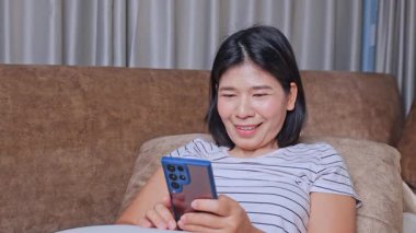 Akıllı telefon kullanan, akıllı telefonuna gülümseyen, aniden sevinçle gülümseyen, sevinçle kutlayan, iyi habere duygusal tepkiler veren, koltukta oturan başarılı genç bir kadın.