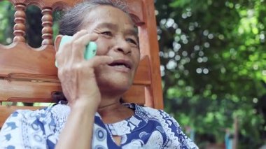 Mutlu Asyalı olgun kadın bahçede mutlu bir şekilde gülümsüyor, ailesiyle cep telefonunda konuşuyor, Tayland 'lı yaşlı büyükanne kablosuz teknoloji ve dijital cihazla dışarıda akıllı bir yaşam tarzının tadını çıkarıyor.