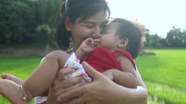 Küçük kız annesiyle oynaşıyor, mutlu sarılıyor ve göbeğini öpüyor. Tayland 'lı genç anne, o parkta gülerken ve mutlu bir şekilde bebeğini kucağına alıp kaldırıyor.