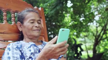 happy asian senior mature woman relaxing and using smartphone, speaking on mobile phone with family, thai asia elderly grandmother enjoy smart outdoor lifestyle with wireless technology device