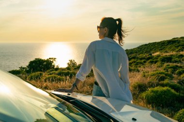 Beyaz kadın deniz kenarına park etmiş arabanın yanında duruyor ve günbatımının tadını çıkarıyor, şehirden kaçıyor.