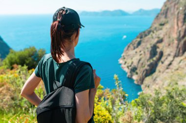 Kadın sırt çantasıyla uçurumda duruyor, okyanus manzarasının tadını çıkarıyor.