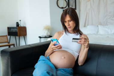 Hamile bir kadın internetten bebek alışverişi yapıyor. Kadın ödemeyi akıllı telefonuyla yapıyor ve evde koltukta otururken plastik kredi kartı kullanıyor.