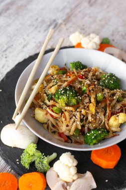 Wok, noodle, sebzeli udon, siyah tabak ve tahta beyaz arka plan. Çiğ sebzeler