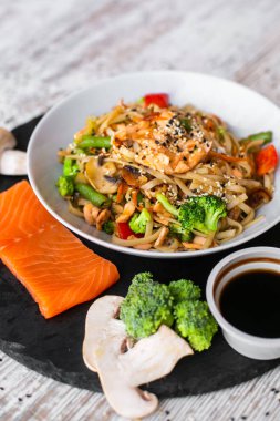 Wok, noodle, somonlu udon, siyah tabak ve ahşap beyaz arka plan. Çiğ sebzeler