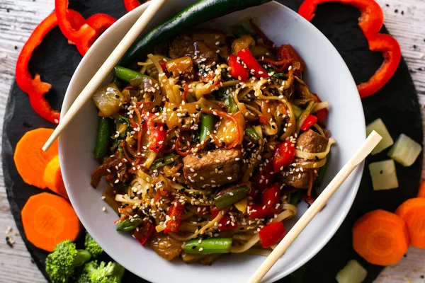 Wok, noodle, domuz eti ve anananaslı udon, siyah tabakta ve tahta beyaz arka planda. Çiğ sebzeler