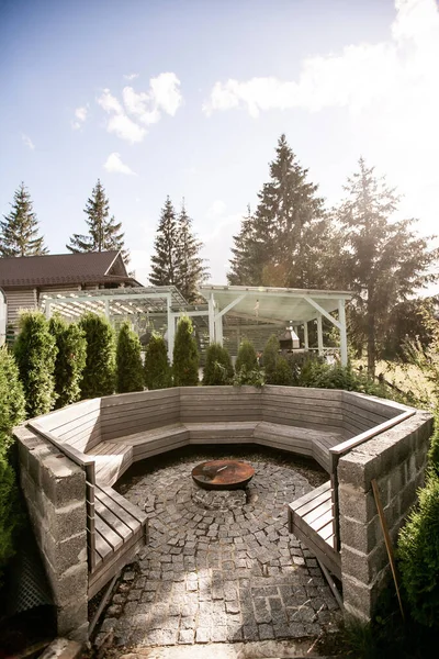Patio design. Patio made of natural stone and composite boards. Round table with a hidden hearth. Benches around the table. Flower beds around