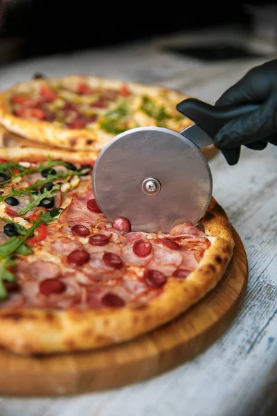 Cutting pizza on slices with special knife. Fresh pizza at background. Hand holding knife.