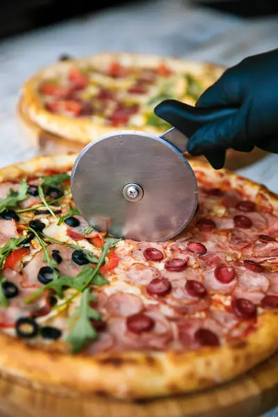 Cutting pizza on slices with special knife. Fresh pizza at background. Hand holding knife.