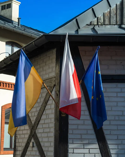 Flags of Ukraine, European Union and Poland next to a brick wall of an old building under a blue sky on a street in the city of Krakow, Polish and European solidarity with Ukraine